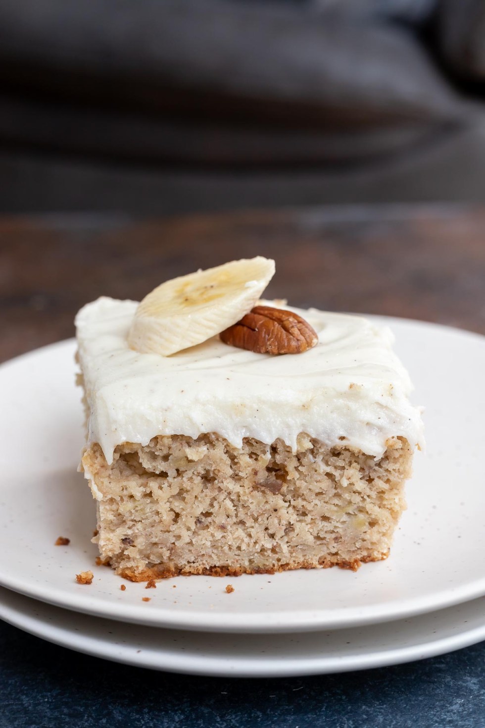 piece of banana cake topped with cream cheese frosting, a banana slice and a pecan. Served on white plate