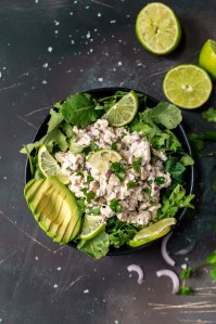 over head photo of cilantro lime chicken, sliced avocado in black bowl on black background, limes on the side