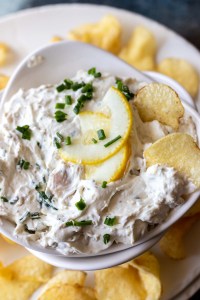 creamy dip topped with lemon slices and chopped chives in white bowl next to potato chips