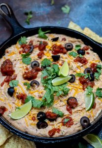 hot bean dip topped with black olives, cilantro, salsa, limes and shredded cheese in cast iron pan