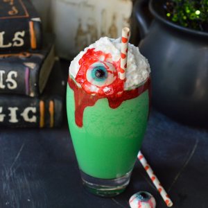 bright green milkshake topped with whipped cream, red sugar gel and gummy eyeball in clear glass