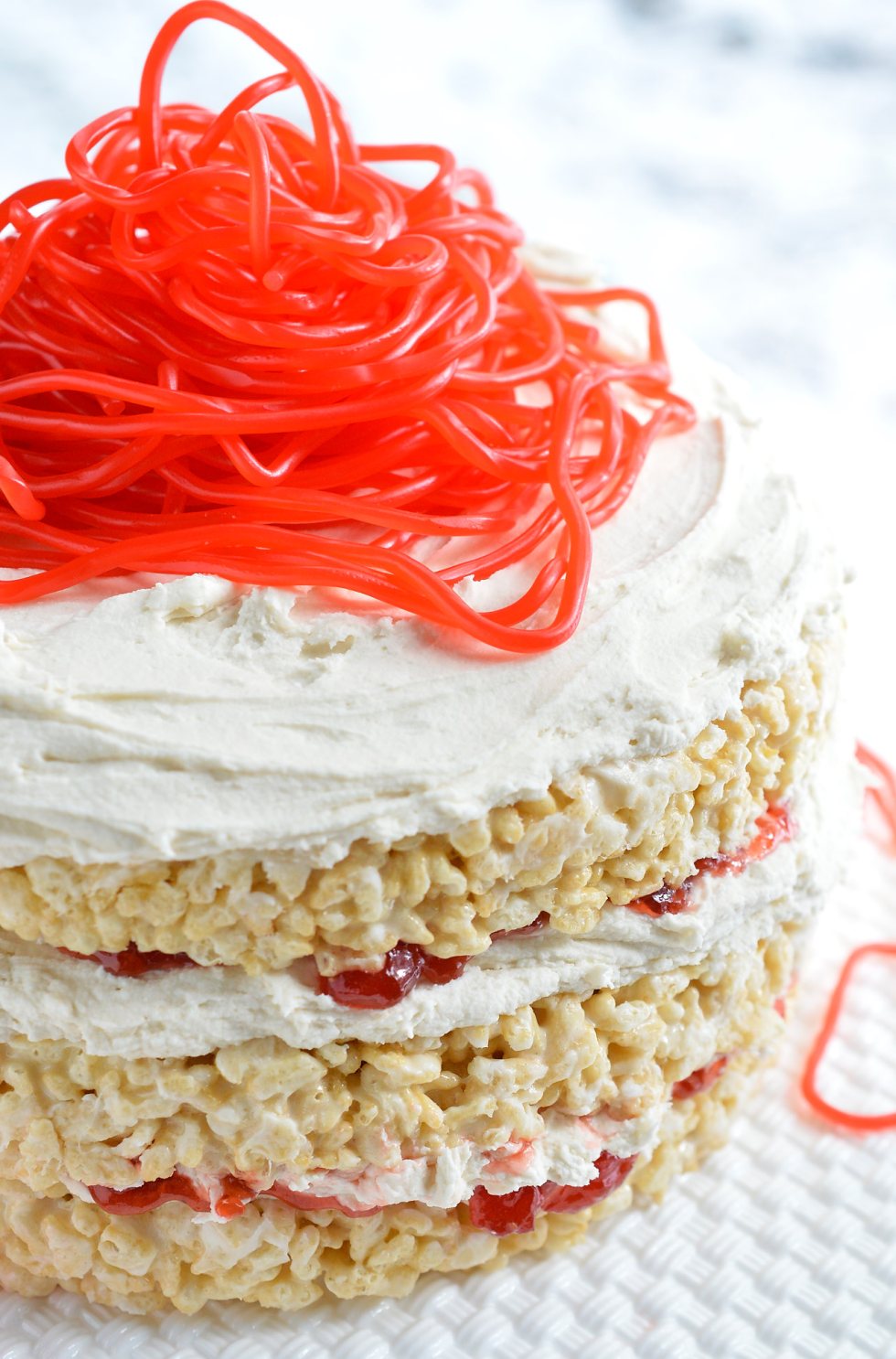 This Rice Crispy Treat Candy Cake is a fun and whimsical dessert recipe. Perfect for Valentine's Day, kids birthdays or for anyone that doesn't like traditional cake. Three layers of rice crispy treats smeared with vanilla butter cream, strawberry jam then topped with strawberry licorice ropes!