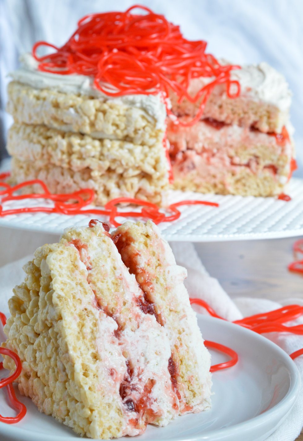 This Rice Crispy Treat Candy Cake is a fun and whimsical dessert recipe. Perfect for Valentine's Day, kids birthdays or for anyone that doesn't like traditional cake. Three layers of rice crispy treats smeared with vanilla butter cream, strawberry jam then topped with strawberry licorice ropes!