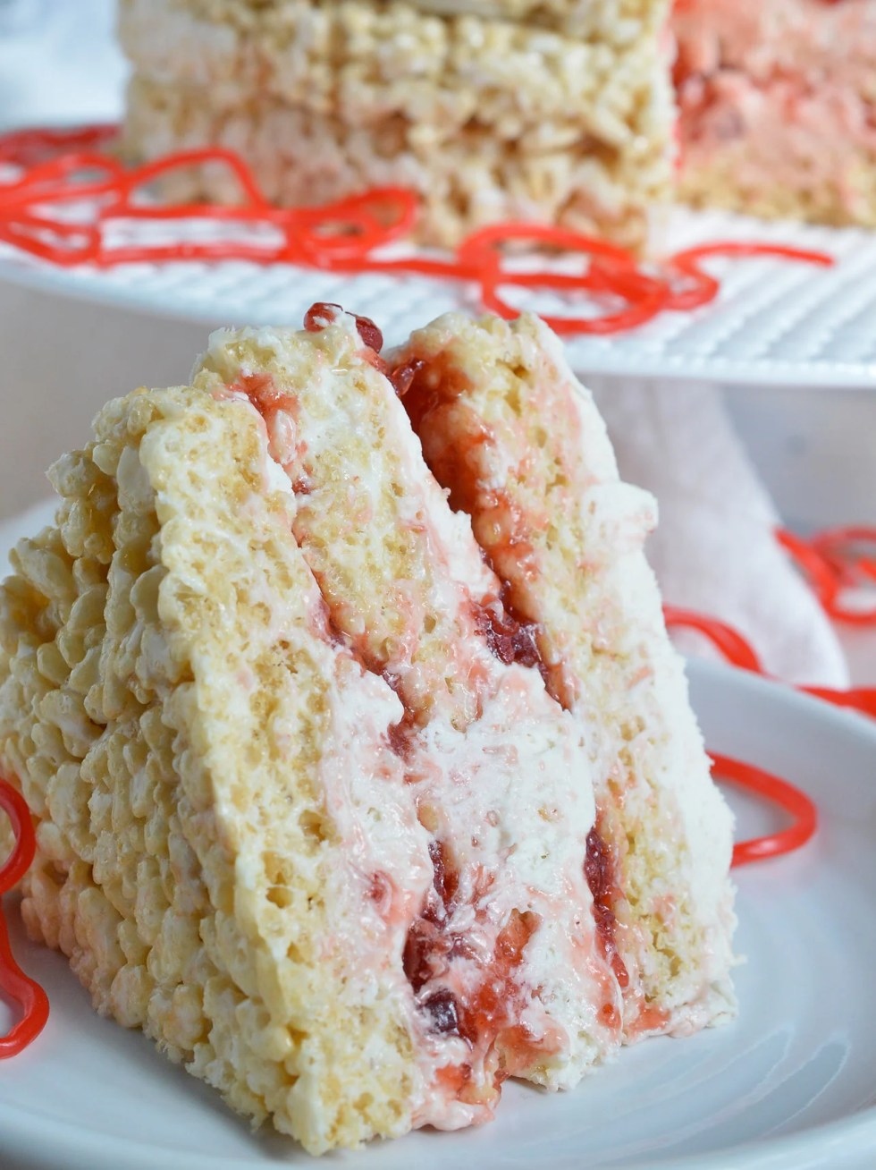 This Rice Crispy Treat Candy Cake is a fun and whimsical dessert recipe. Perfect for Valentine's Day, kids birthdays or for anyone that doesn't like traditional cake. Three layers of rice crispy treats smeared with vanilla butter cream, strawberry jam then topped with strawberry licorice ropes!